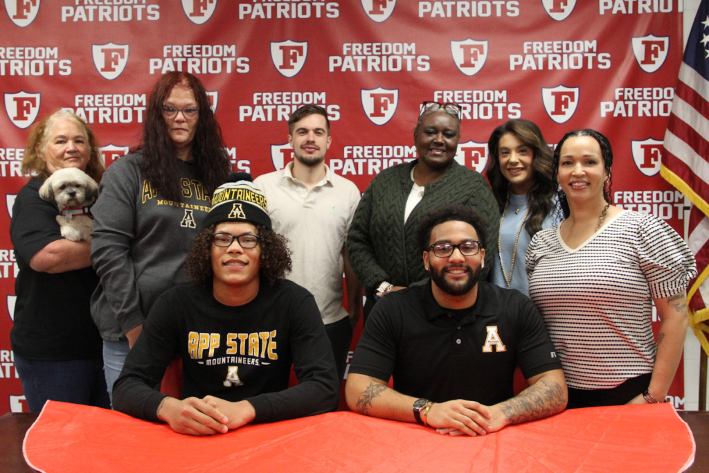 Nathaniel and Jacorian pose with their loved ones in front of a red Freedom High School Patriots banner. 